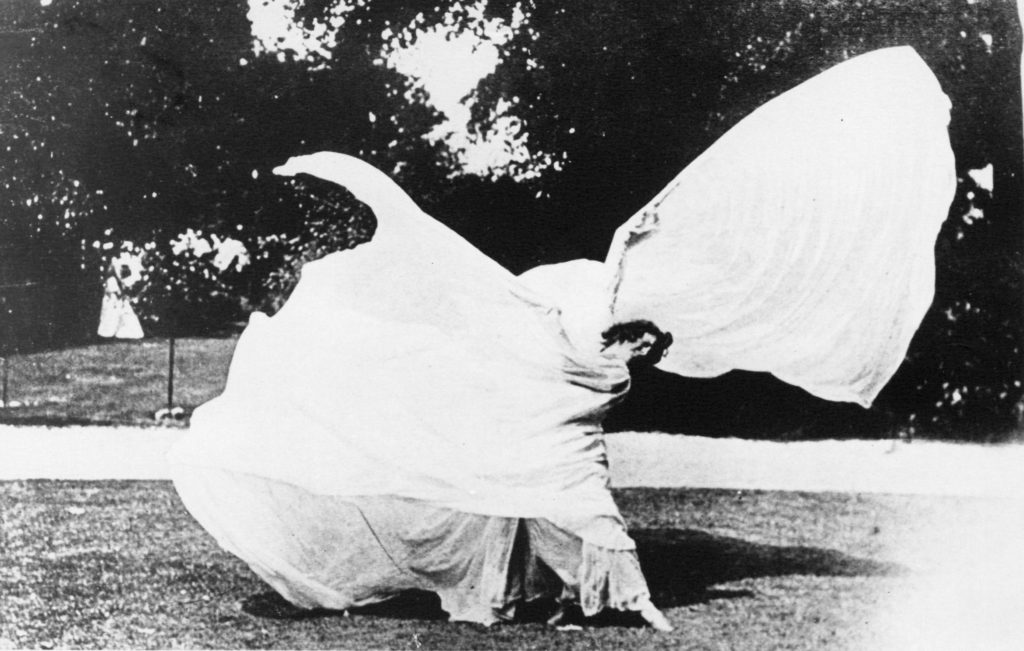 Black and white image of Loie Fuller dancing in a field. She wears her flowing dress with large wing-sleeves on either side and twists her body around so that the fabric billows in a spiral. 