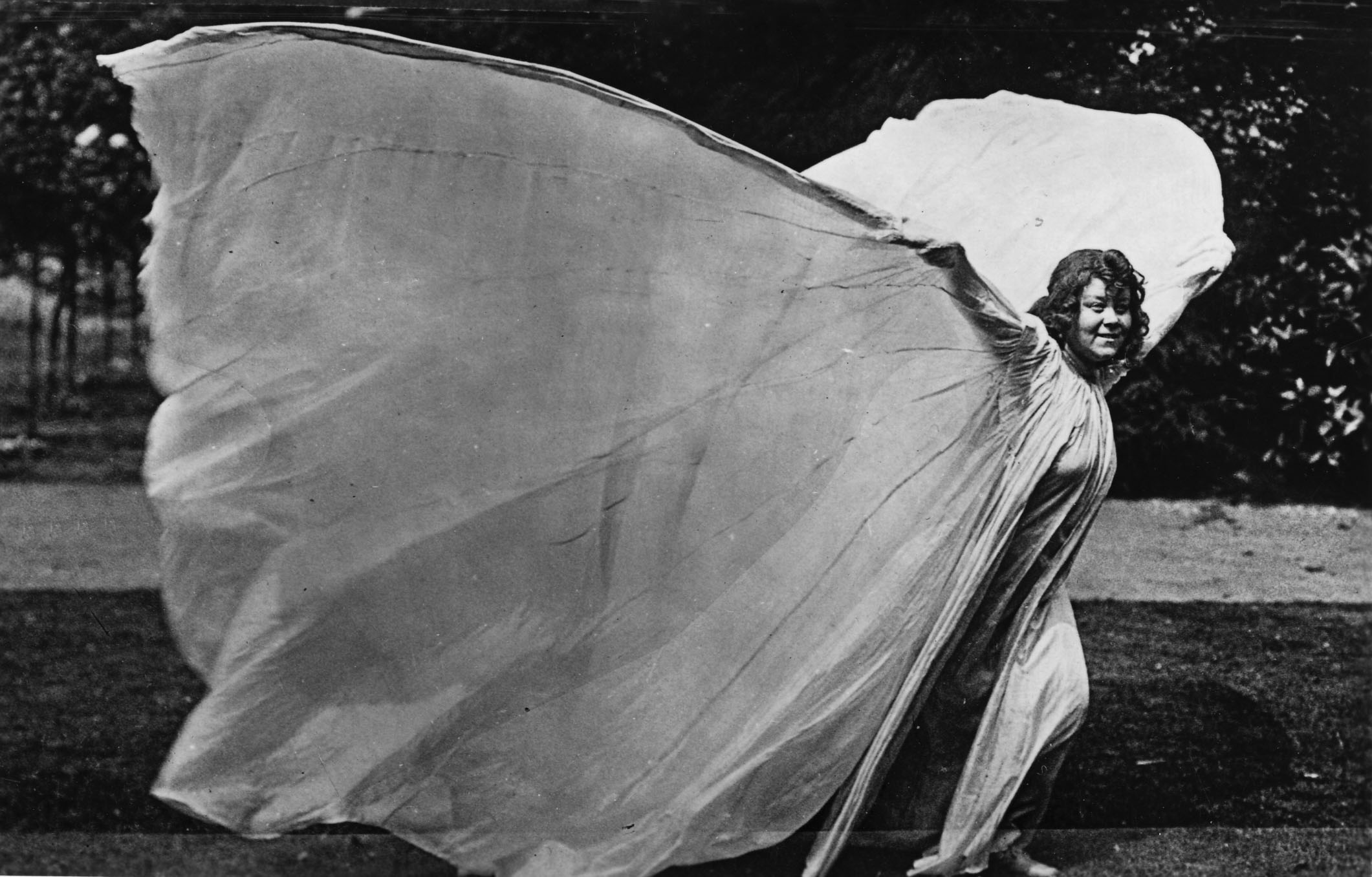 Black and white photo of Loie Fuller in a field. She holds her arms out to support large, floor-length fabric wings on either side of her.