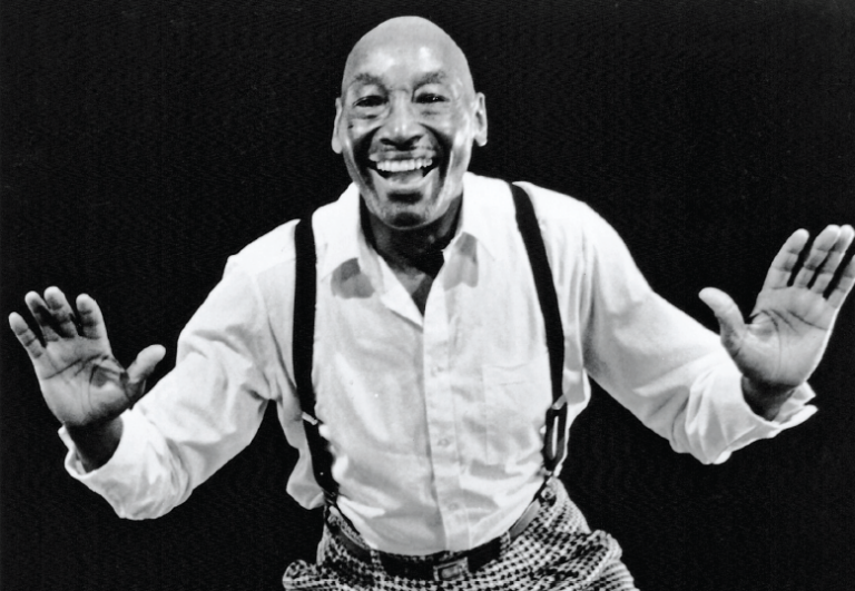 A black and white photo. Frankie Manning, in a white button-up with suspenders and gray pants, raises his hands playfully as he tap dances. He is shown waist-up.