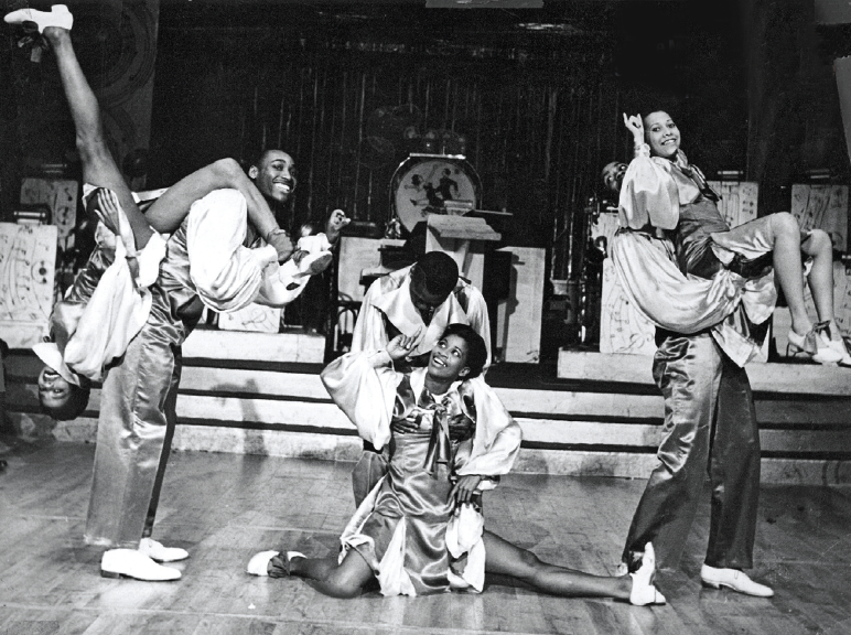 A black and white photo. Onstage, three pairs of male and female partners dance Lindy hop in various poses.