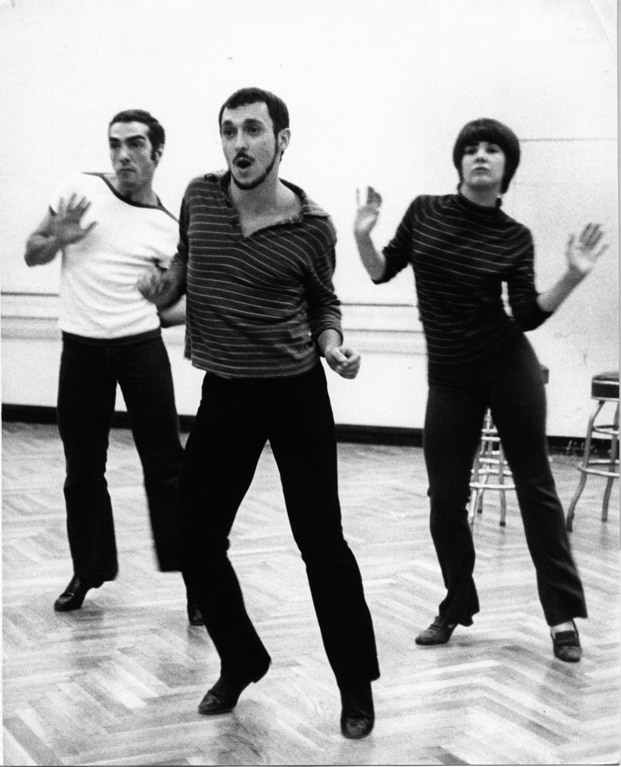Michael Bennett, Bob Avian and Margo Sappington stand in a triangle in the middle of a dance studio. Their hips are jutted to the site, caught mid-bounce by the camera. Photo is black and white.