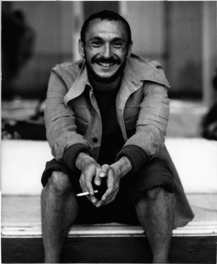 Photo of Michael Bannet sitting on a step, with a cigarette in his clasped hands. He laughs into the camera, wearing a long coat and knee-length shorts. Black and white photo.