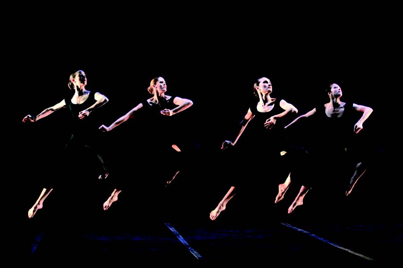A group of four dancers arranged in a line on a dark stage all complete matching stag jumps with their arms lifted across their body. All dancers wear dark calf-length bottoms and tank tops.