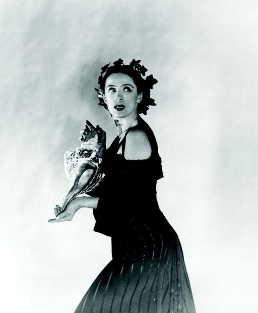 Black and white photo of Martha Graham. She stands with her body faced to the side with her head turned toward the camera. She wears a long, dark dress with an off-the-shoulder top and an ornate flower crown over her head.