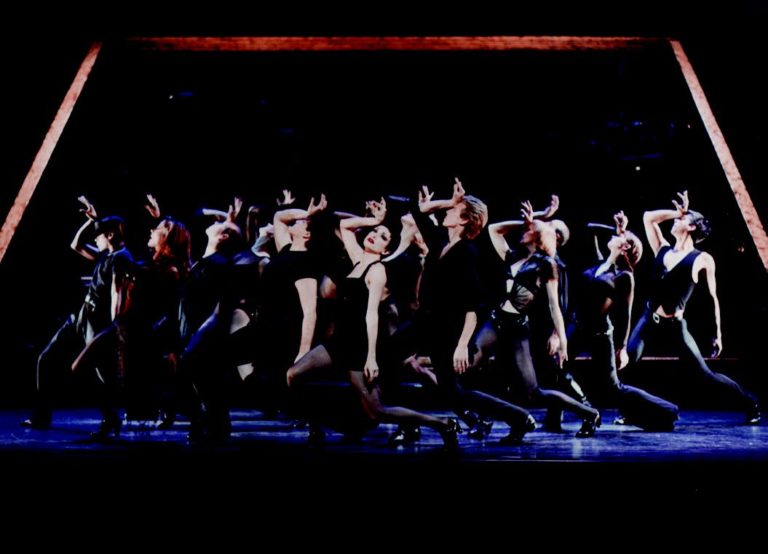 A group of dancers in varied black clothing stand in a cluster with their left hands raised to their foreheads.