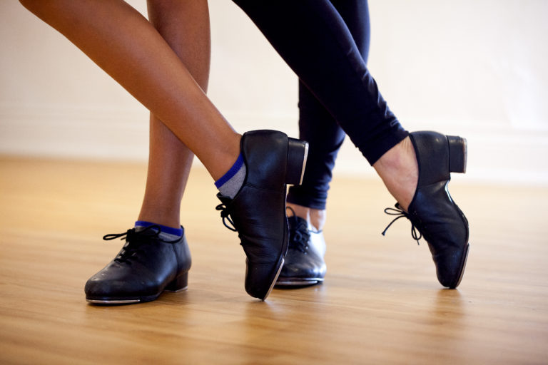 From the ankles down, two dancers in black tap shoes cross one foot over the other