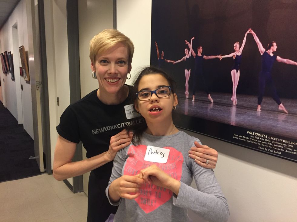 A woman with short blonde hair wearing a New York City Ballet shirt poses behind Audrey, who wears glasses and a name tag.
