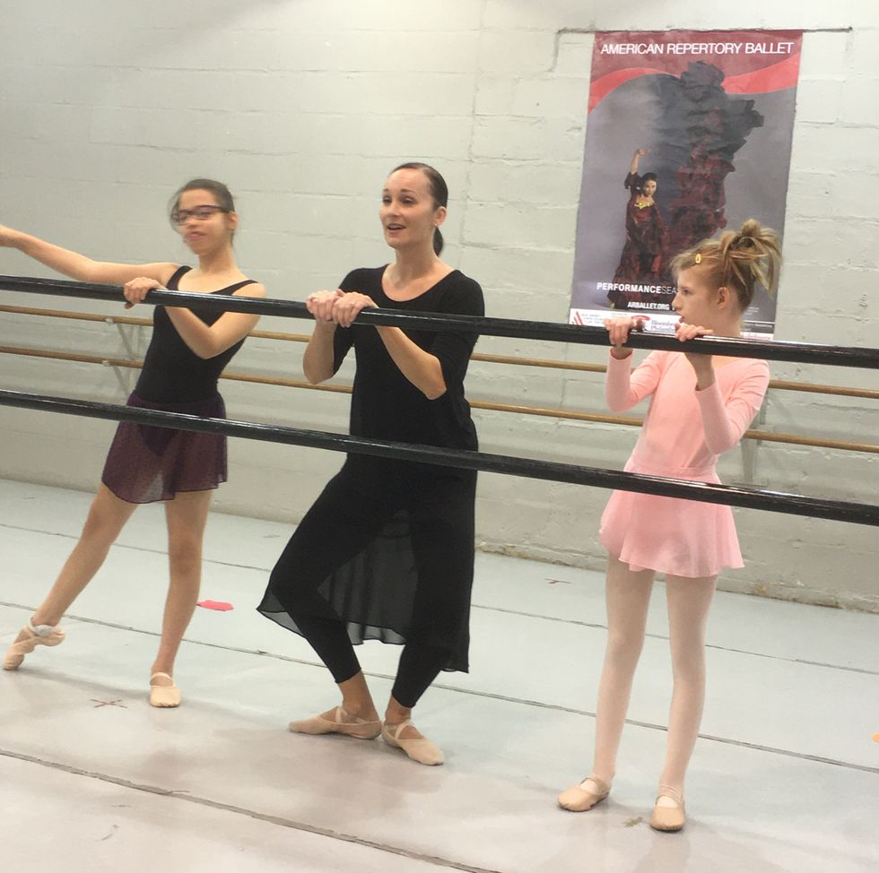 Valerie Amiss stands at the barre in a pliu00e9 in first position between two young ballet students.
