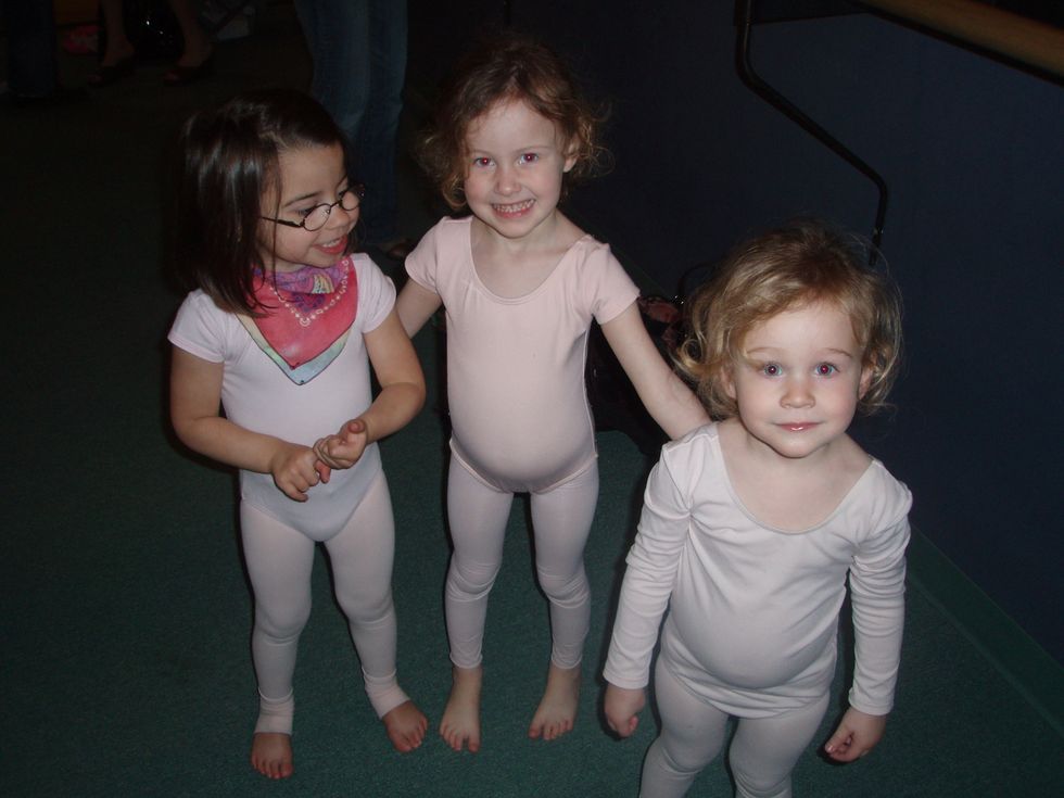 Three 4-year-old dancers in pink leotards and tights stand in a row and smile at the camera. Audrey wears glasses and a bandana around her neck.