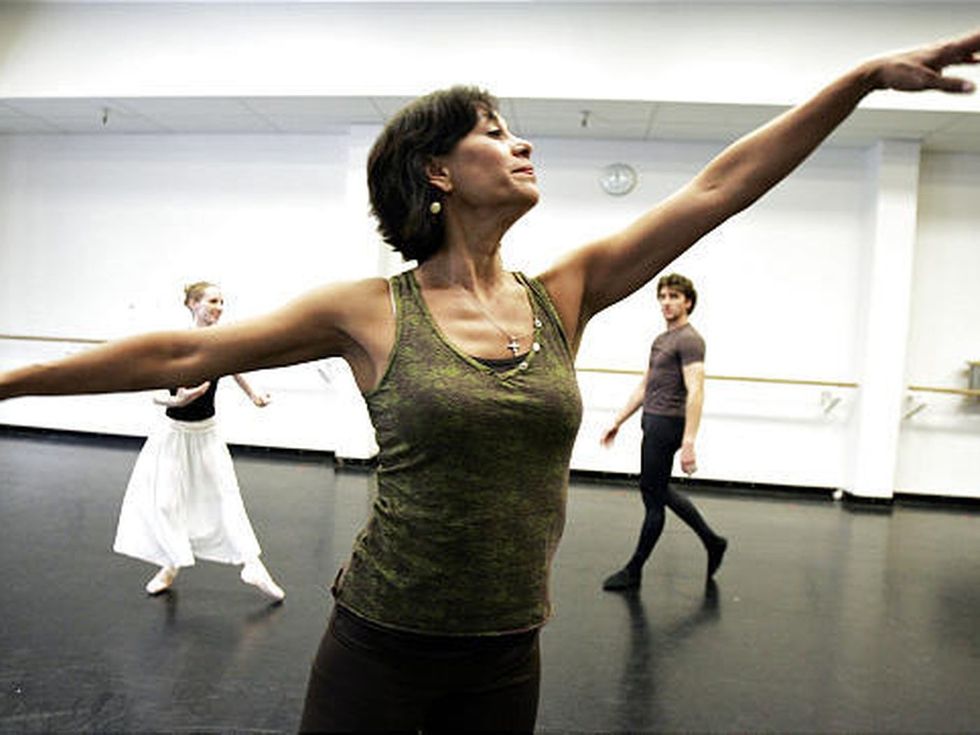 Evelyn Cisneros-Legate demonstrates first arabesque arms, limbs stretching beyond the edge of the photo, as a male and female dancer in rehearsal clothes watch from the back of the studio.
