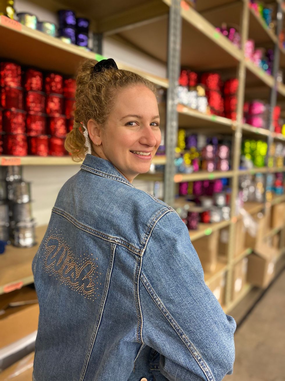 A woman stands in front of a wall of shelves covered with rolls of colorful sequins. She wears a denim jacket that says "wish" on the back in rhinestones