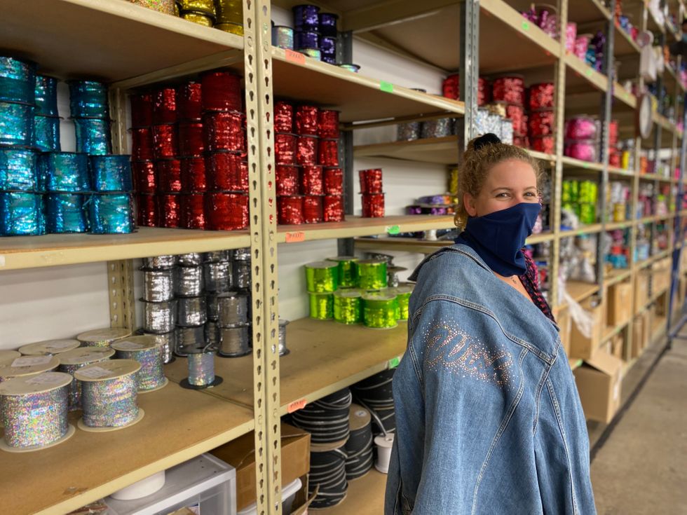 A young woman wearing a mask and a denim jacket with "wish" bedazzled on the back stands in front of a wall of shelves full of sequins