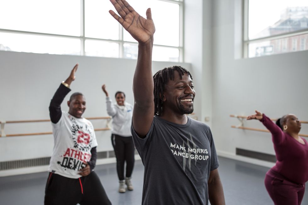 A Black male dance teacher raises his right arm overhead, smiling, while adult students follow along.