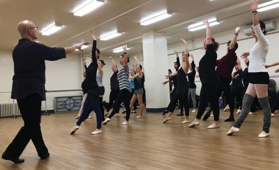 Finis Jhung, wearing black pants, a black shirt and glasses, speaks to a large group of adult students as they execute a tendu combination in center.
