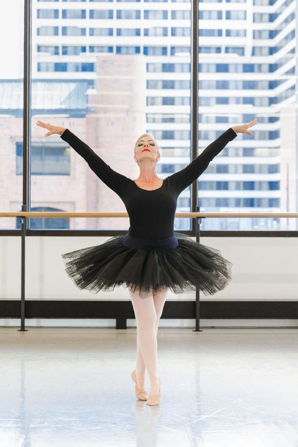 A middle aged woman in a long-sleeved black leotard, black tutu, pink tights and ballet slipper, poses dramatically in sus-sous, her arms in a V shape.