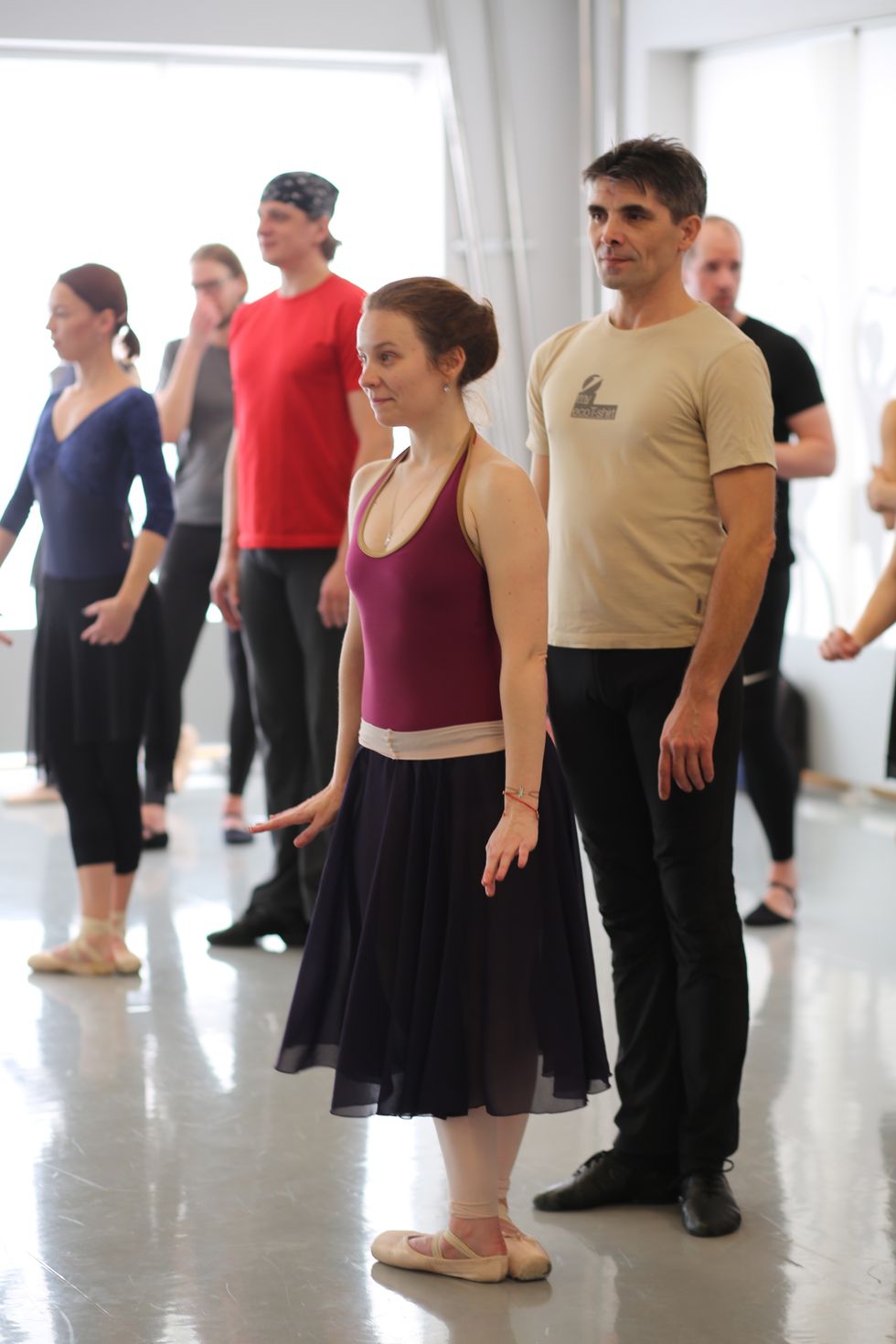 During a pas de deux class, a woman in a burgundy leotard, black skirt and pointe shoes stands in fifth position while her partner, in a beige T-shirt and black pants, stands behind her in a loose first position.