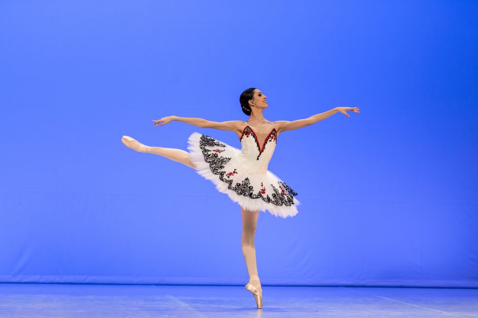Carolina Pires does a piquu00e9 first arabesque on pointe on her left leg. She smiles brightly, wearing a white tutu with black and red trim, pink tights and pointe shoes. Her dark brown hair is pulled into a low bun.