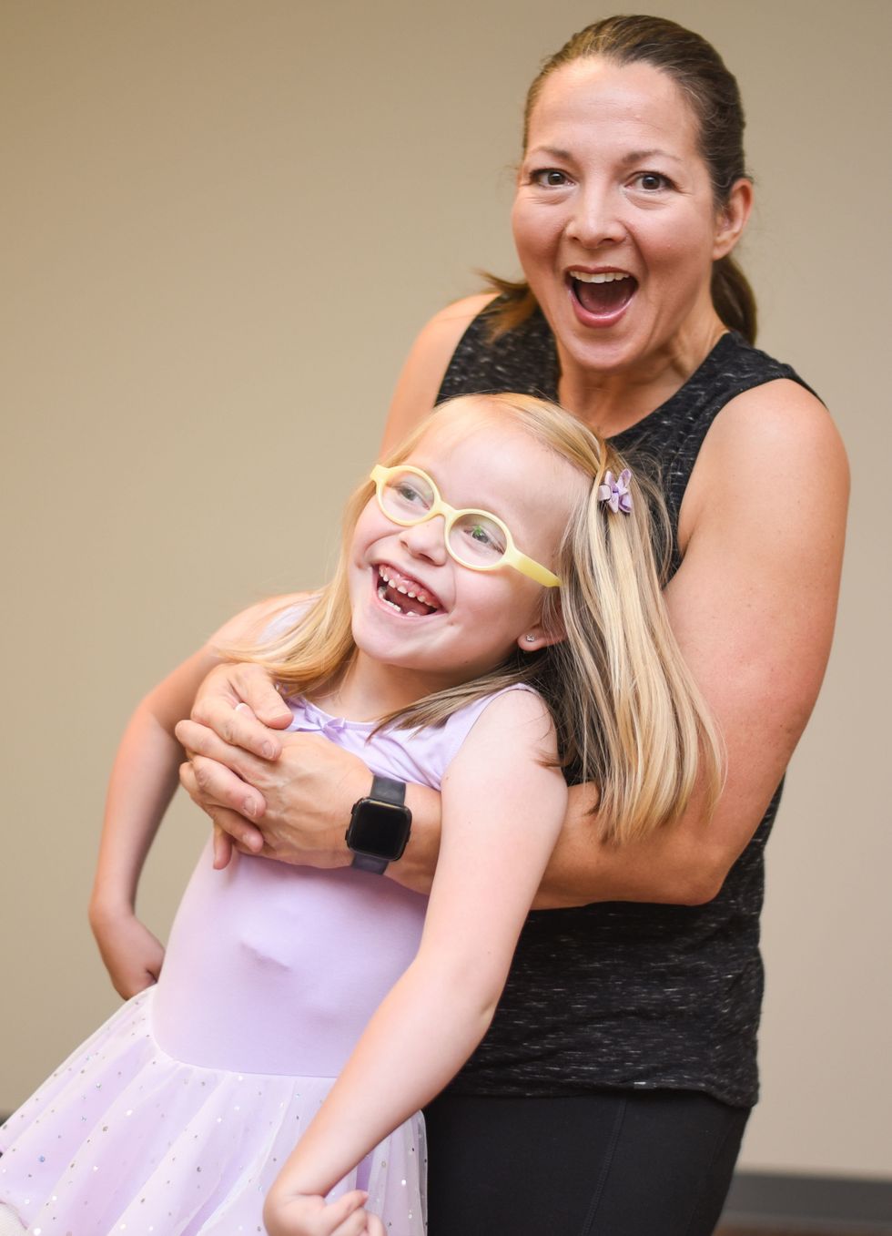 Kim Black grins at the camera, holding on to a young girl in a sparkly purple dress and yellow glasses who is also grinning