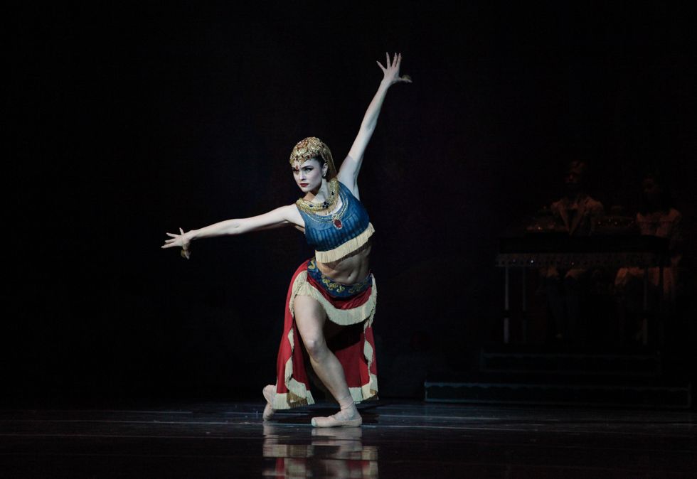 Caitlyn McAvoy, wearing a blue, gold and red costume and gold headress for the Nutcracker's Arabian dance, lunges deeply in fourth position with her arms spread wide.