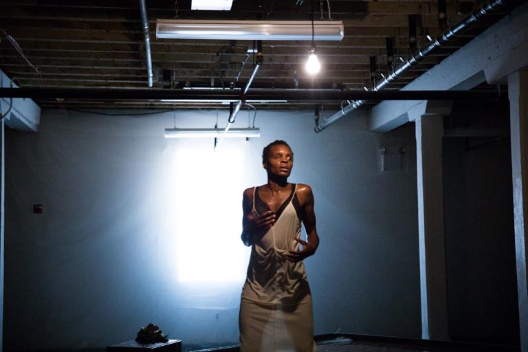 Okpokwasili, a tall Black woman, stands in a dark, scarce loft-like space, with a bright light on her. She wears a white slip, and appears to be wet