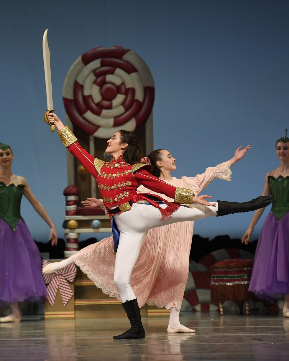 A performance image of the Nutcracker in front of Clara. Each are doing an arabesque in an opposite direction of the other.