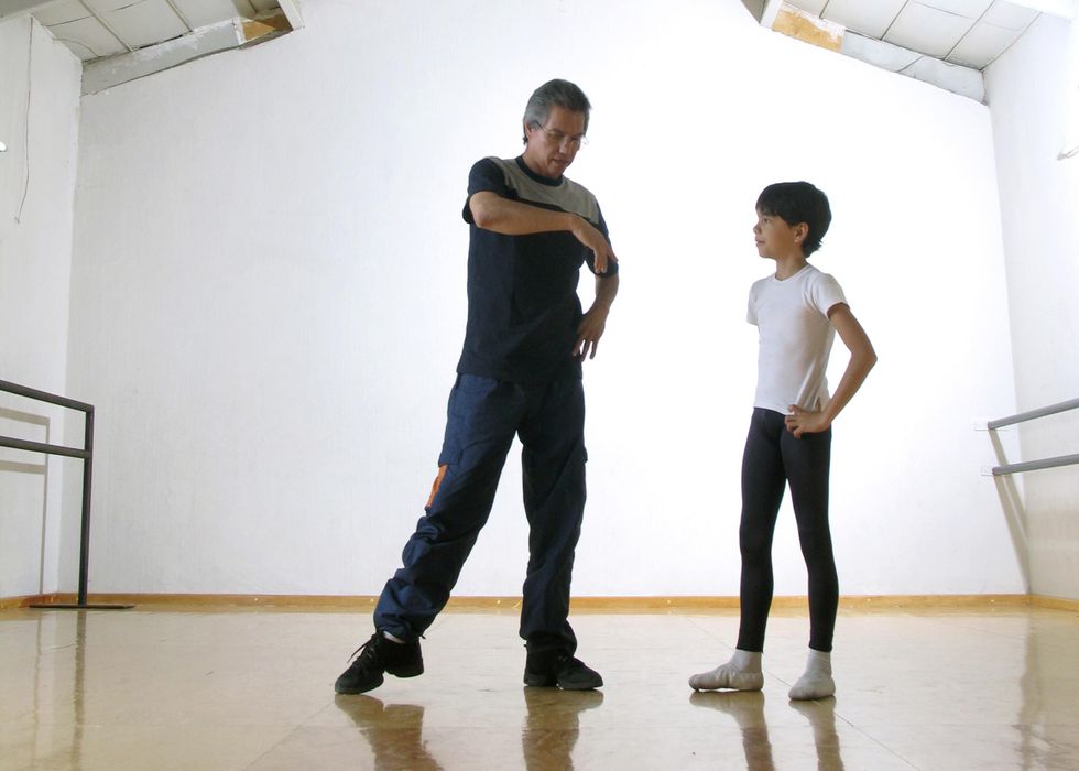 In a sunny studio, an older man in a blue and gray T-shirt and blue workout pants stands on his left leg with his right in a relaxed tendu. He holds his left arm on his hip and moves his right arm across his body while a young boy in a white T-shirt and black tights stands to his left and watches.