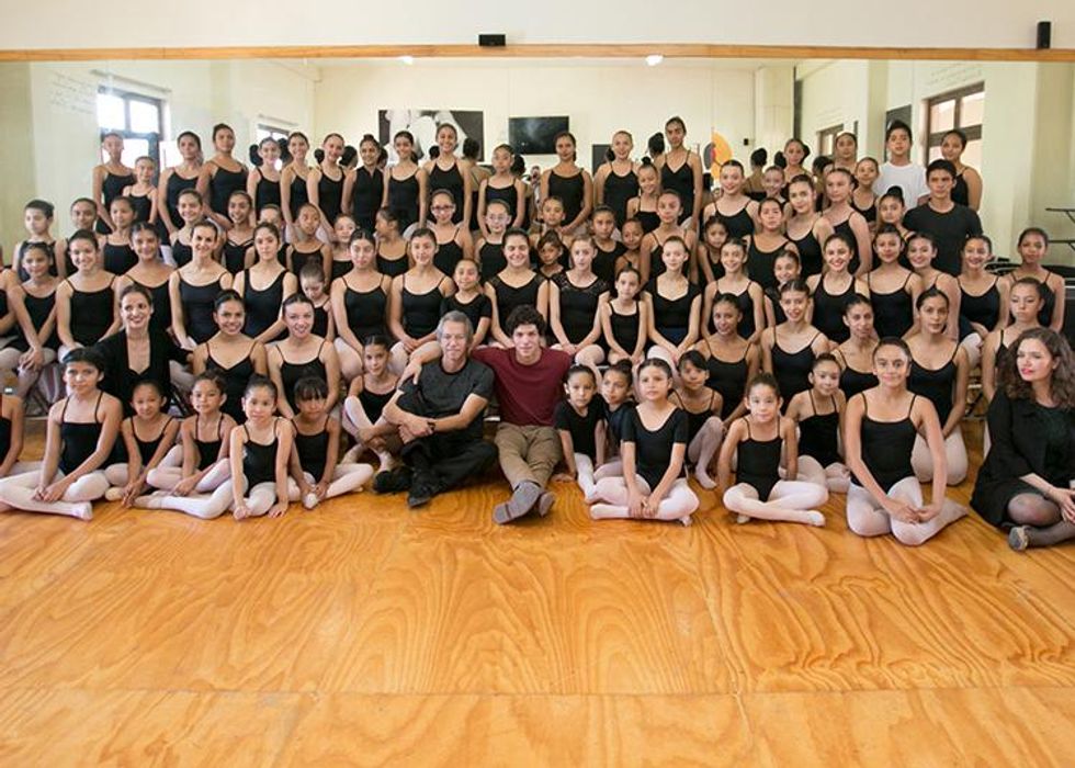 A large group of ballet students  in black leotards and pink tights take a group photo. In the middle, sitting on the floor, sit an older man in a gray T-shirt and black workout pants and to his right, a young man in a burgundy long-sleeved shirt and khaki pants.