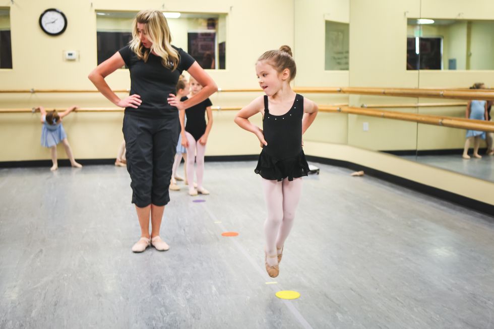 A dance teacher leads a group of young girls in an exercise, they have their hands on their hips and look at themselves in the mirror.