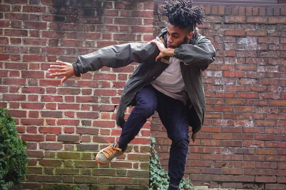 Danzel Thompson-Stout, wearing jeans, sneakers and a green jacket, dances in front of a brick wall