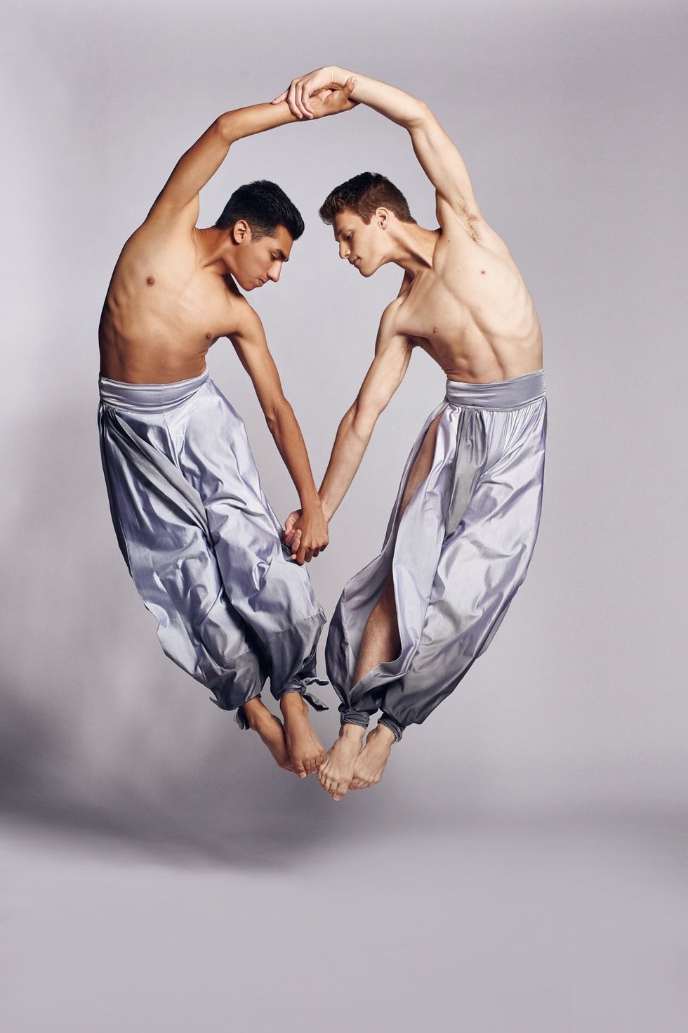 Two male dancers leap into the air, holding handsu2014one above their heads and one above. They wear shiny silver pants, with their heads and feet touching and their hips leaning away.