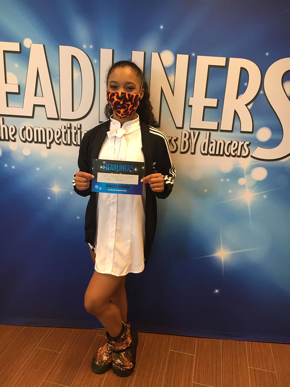 A young masked dancer standing against a Headliners backdrop holds a certificate that says "Headliners Judge's Award"
