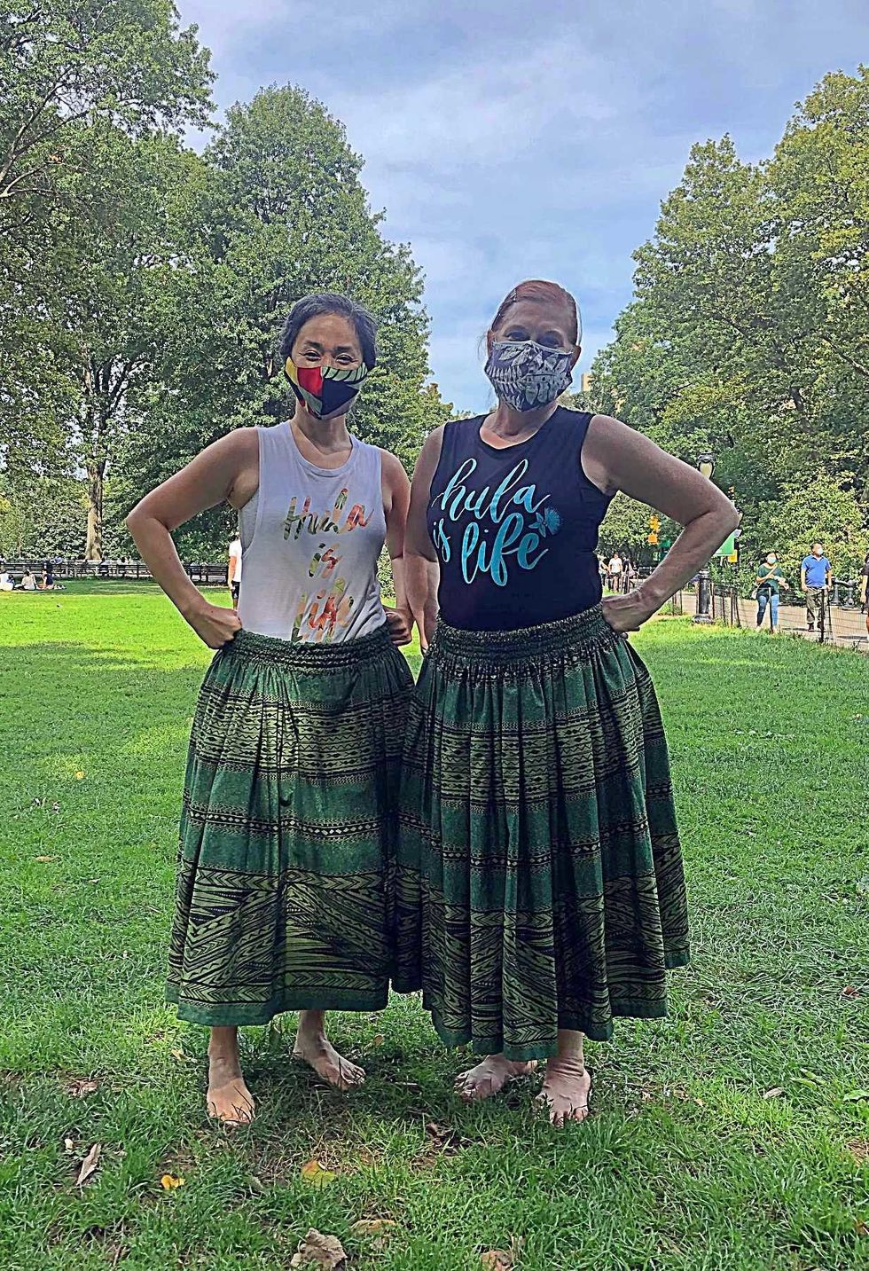 Two women in green skirts, masks, and shirts that say "hula is life," stand in a grassy area in the park