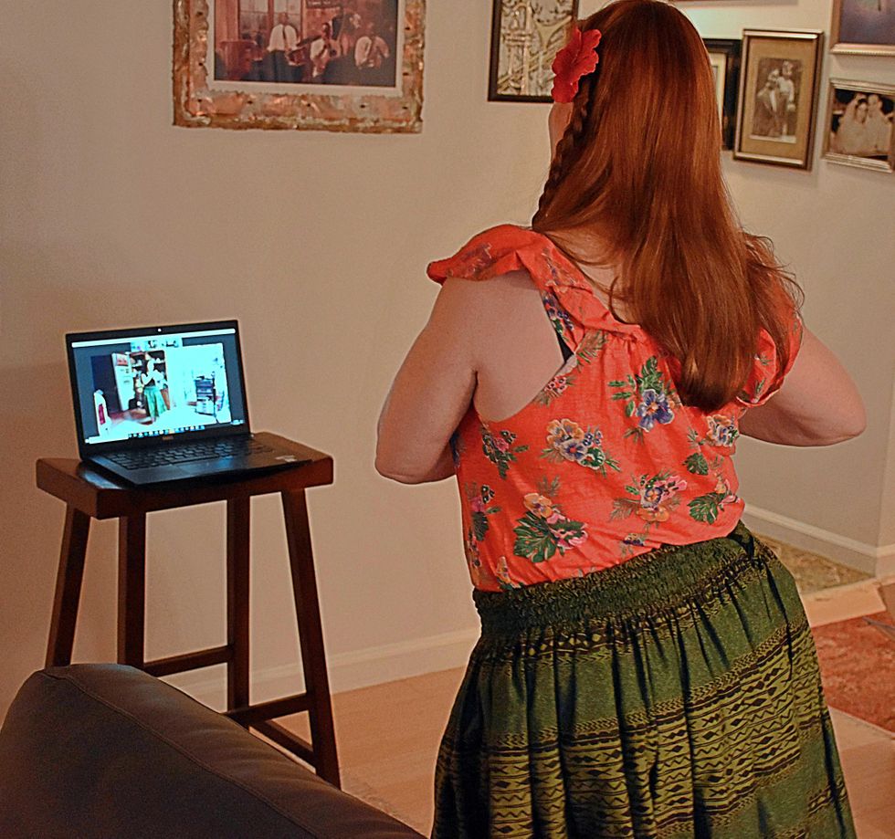 Ruden, wearing a bright red floral top and a green skirt, dances faces her computer which sits atop a stool