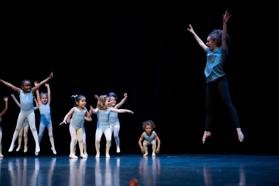 Bailes, in a blue T-shit and black leggings, jumps high in the air with her arms up and her tows pointed. To her left, a group of little girls wearing blue leotards and pink tights try to copy her.