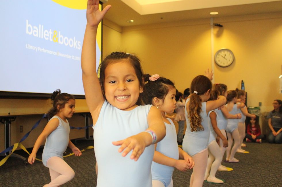 A little girl in a light blue leotard gives a big smile to the camera with her right arm lifted up high and her left arm extended out in front of her.