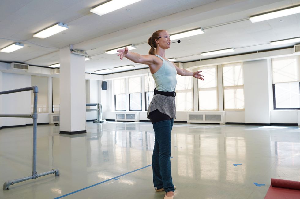 Deanna Doyle, wearing a microphone and a light blue tank top, demonstrates second position arms in an empty classroom