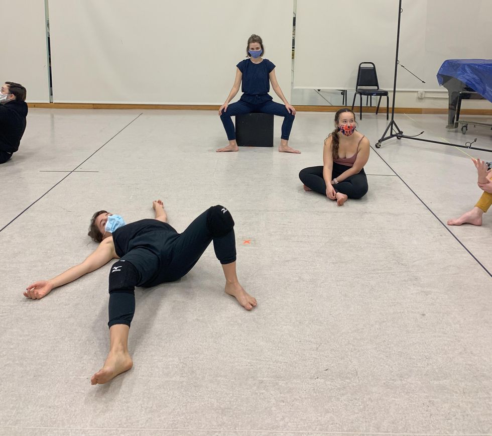 Masked dancers spread out across a studio sit or lay on the floor during a break