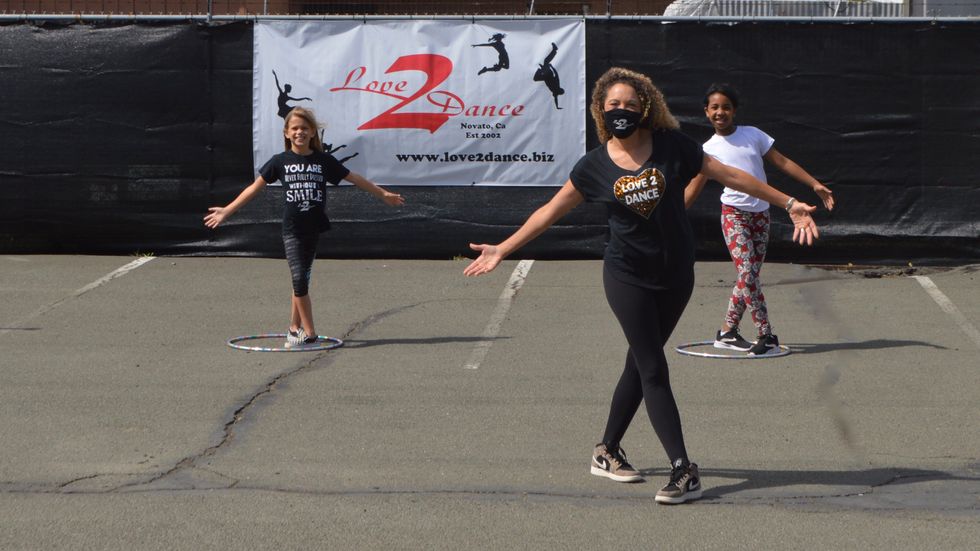 A teacher, wearing a mask, and two young students pose in a parking lot. There's a "Love2Dance" sign in the background, and students are spaced out with hula hoops.