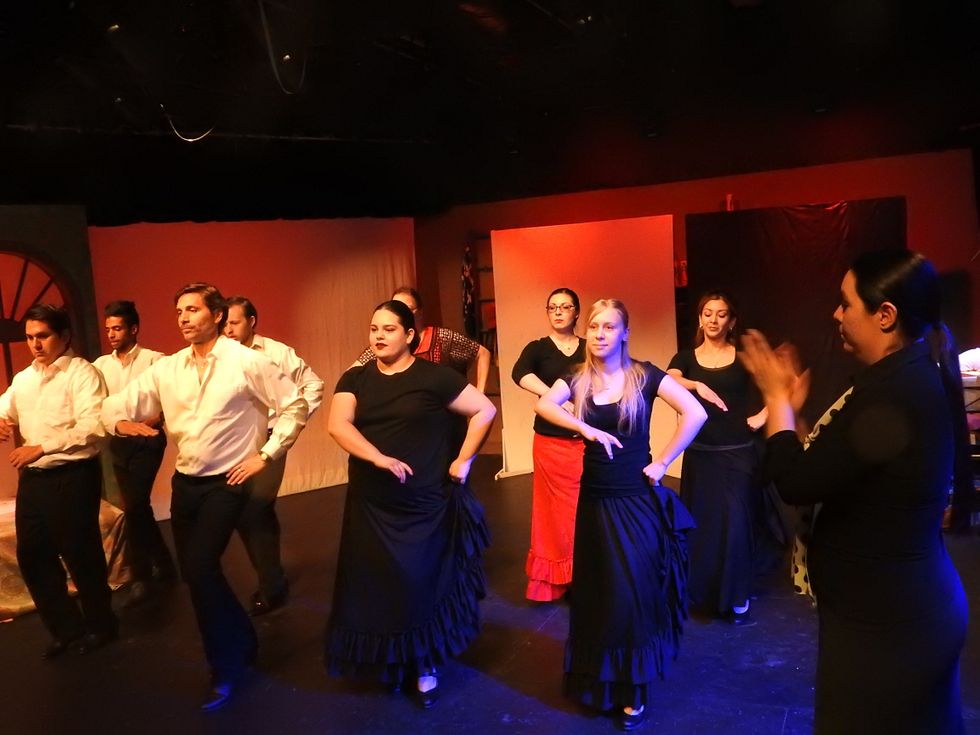 A group of men in white shirts and black pants, and women in black shirts and long skirts, take a flamenco class. Their female teacher stands to the right and claps out the rhythm.