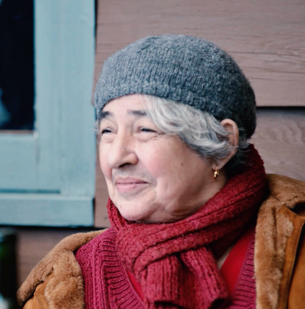 Aileen Passloff, gray and white hair tucked under a grey knit beanie, gives a closed mouth smile as she looks at something off camera.