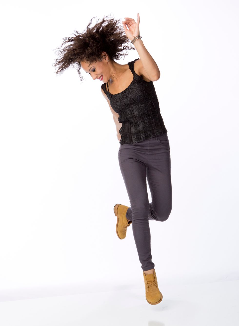 Ayodele Casel hops on one foot against a white backdrop. She is wearing tan tap boots, gray jeans and a black tank.