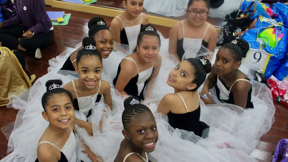 A group of young girls sit on the floor and smile at the camera, wearing white tutus