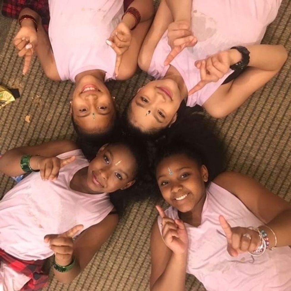 Four girls wearing pink shirts lay on the ground, smiling, with their heads together, make #1 signs with their fingers