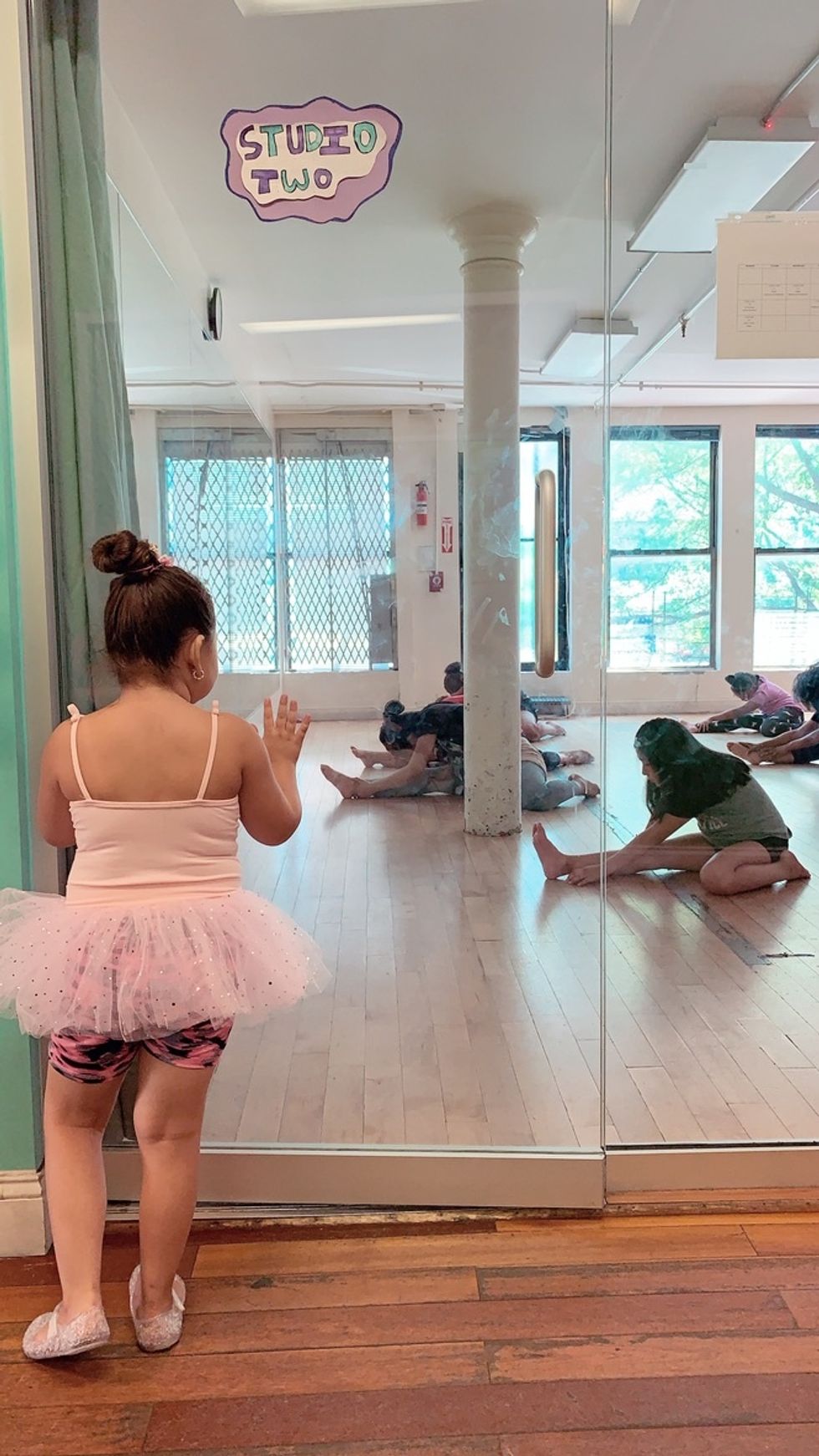 A young girl in a tutu looks through a glass door at a class in session, with students stretching on the floor