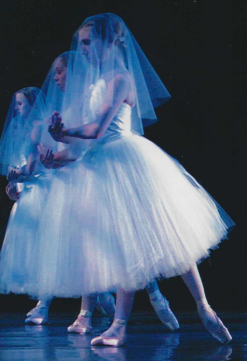 A young ballerina, wearing a white romantic tutu and veil, stands in tendu derriu00e9re with her arms crossed in front of her, palms up. A row of similarly costumed women stand directly behind her in the same pose.