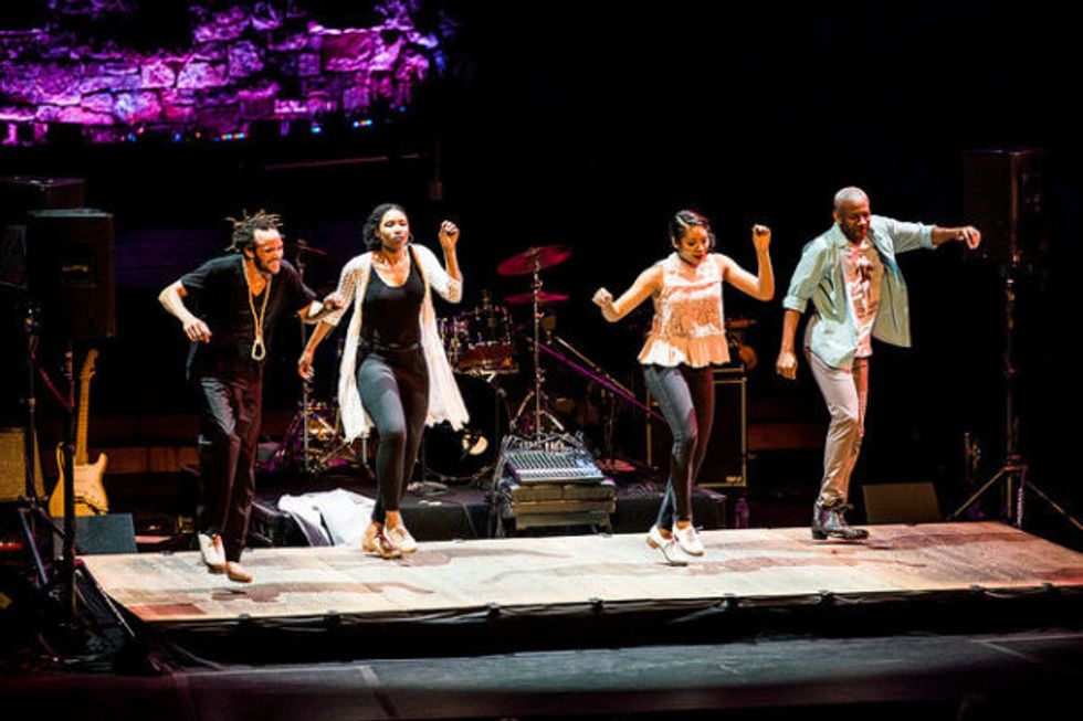 Watson tap dances with three other dancers on raised platform in front of a drum set and some other instruments