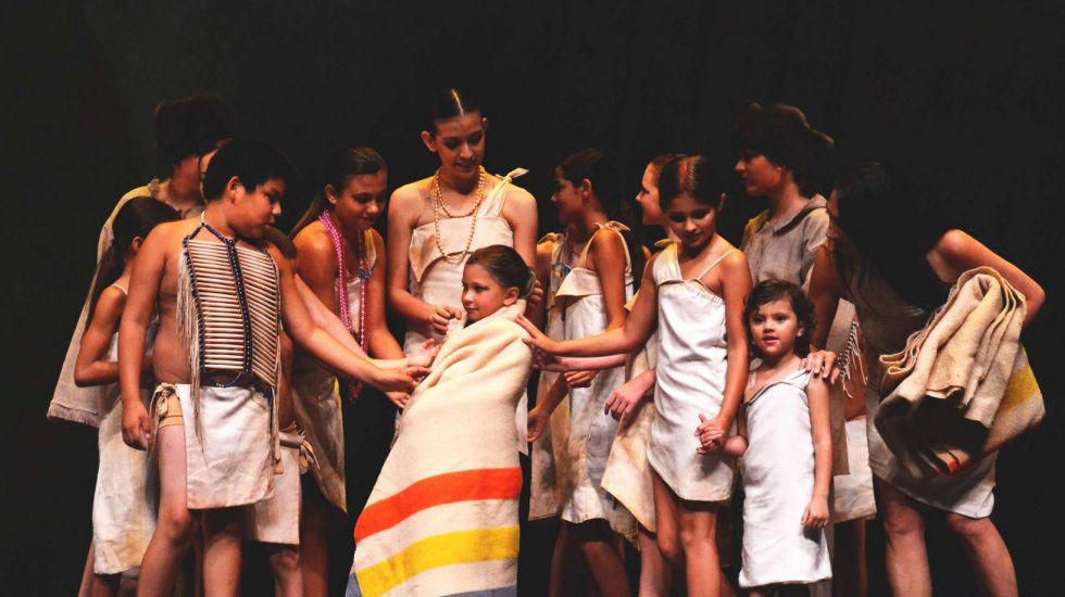 Around 12 young dancers in Osage attire huddle together onstage, smiling at one another