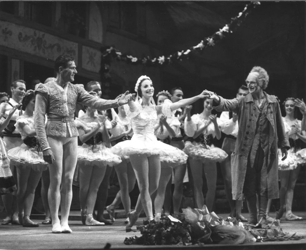 I this black and white photo, a ballerina in a white tutu takes a bow on a crowded stage.