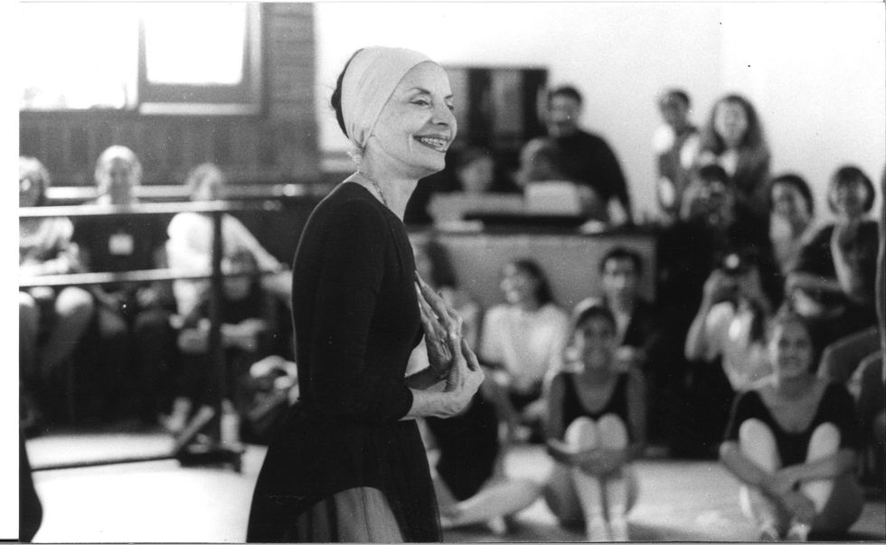 A ballet teacher demonstrates to students in a studio. She wears a head scarf, black leotard, tights and skirt.