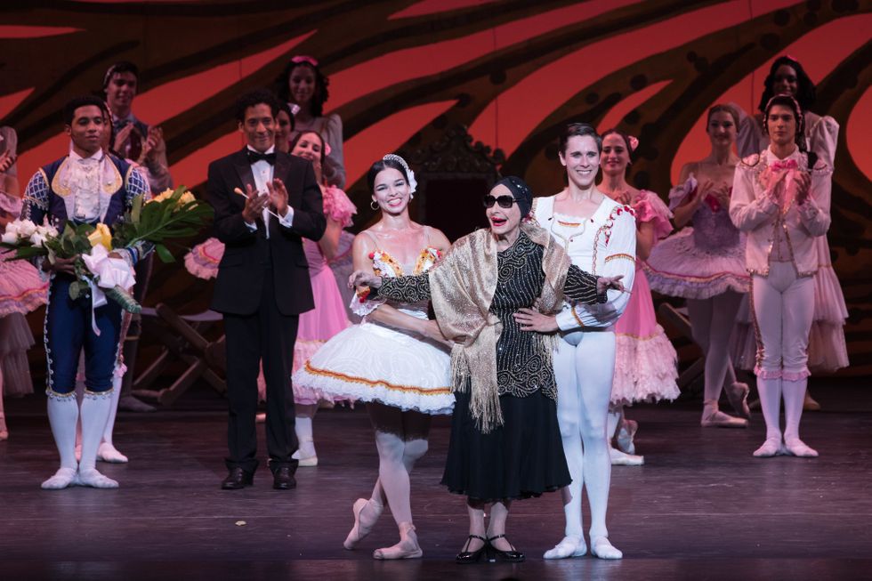 A bakkerina in a tutu, an onlder woman in sunglasses and a dark dress, and a male dancer in white tights and a white tunic, take a bow onstage.